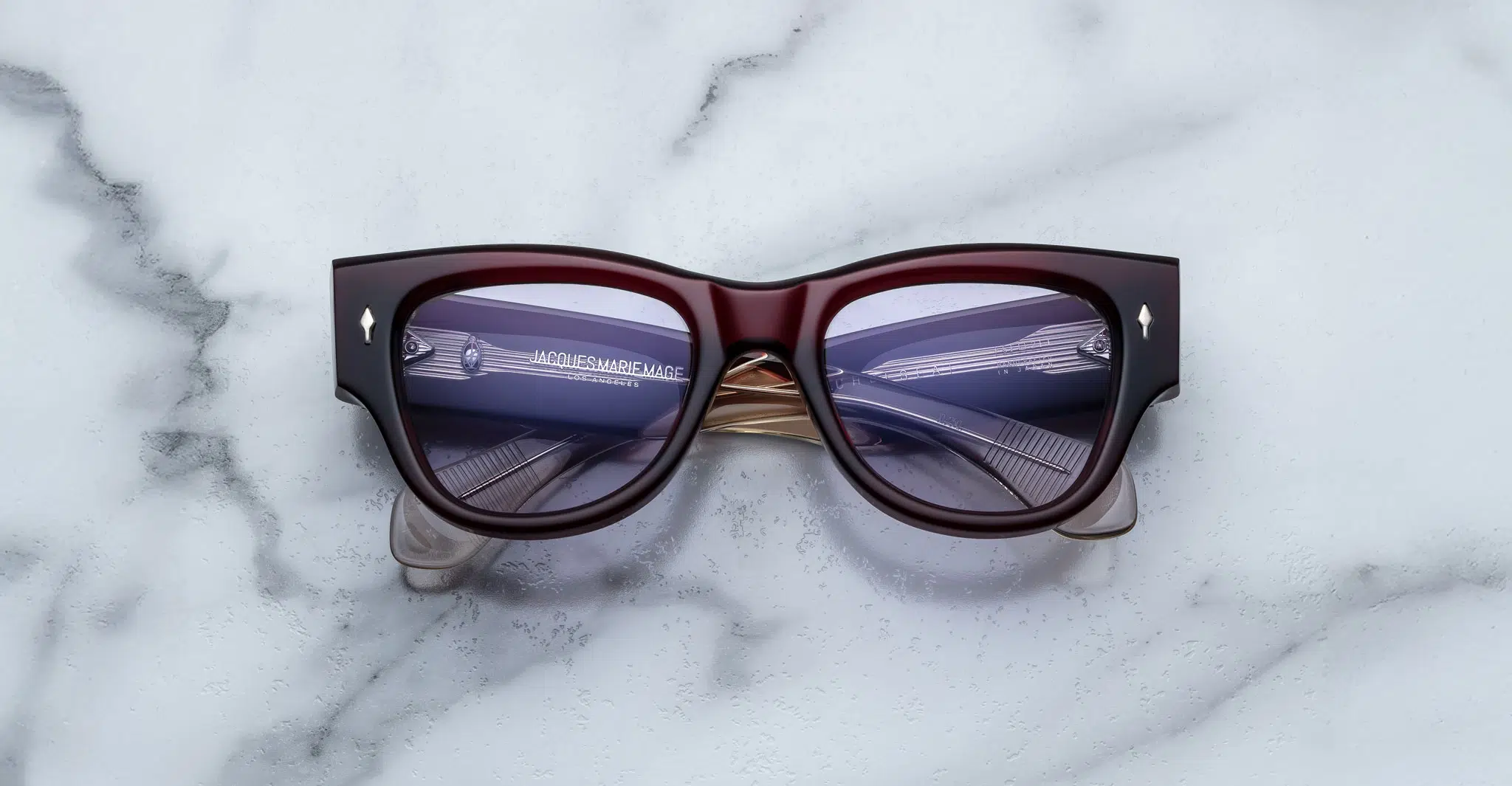 A pair of Jacques Marie Mage Chelsea cherry red, thick-framed sunglasses with light purple lenses rests on a white marble surface with grey veins, with faint brand details visible on the inside of the left lens.
