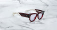 A pair of Jacques Marie Mage Chelsea cherry red, thick-framed sunglasses with light purple lenses sits on a white marble surface with grey veins