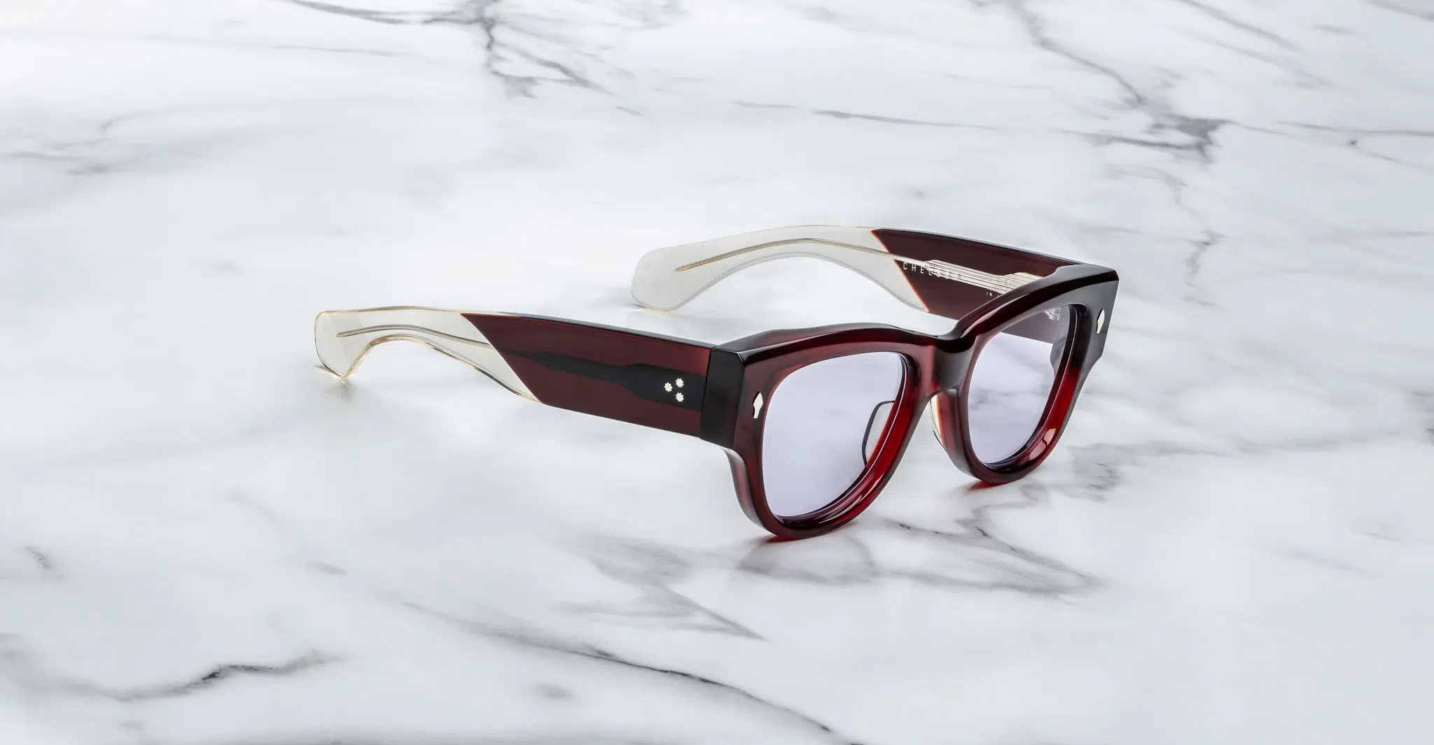 A pair of Jacques Marie Mage Chelsea cherry red, thick-framed sunglasses with light purple lenses sits on a white marble surface with grey veins