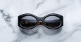 A pair of Jacques Marie Mage Gia black, thick-framed sunglasses with grey lenses rests on a white marble surface with grey veins, with faint brand details visible on the inside of the left lens.