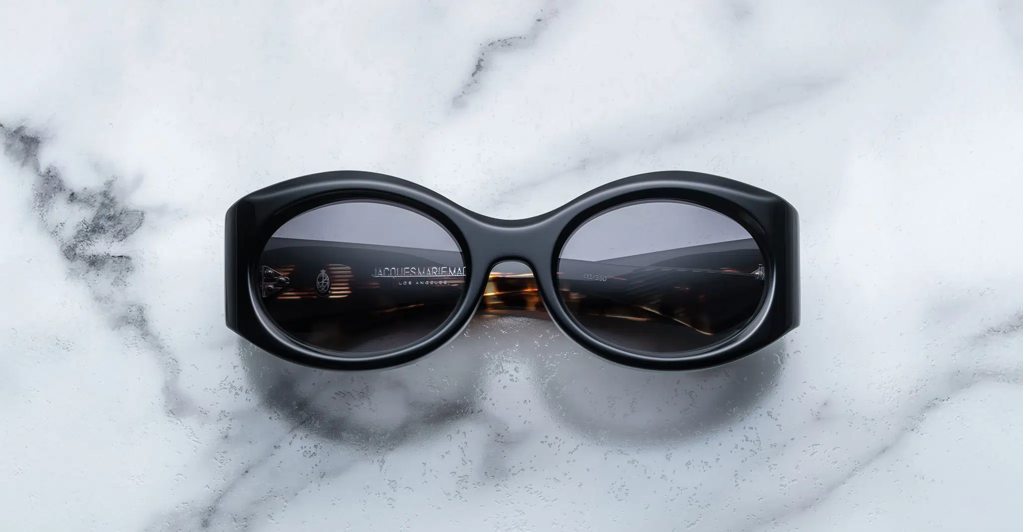 A pair of Jacques Marie Mage Gia black, thick-framed sunglasses with grey lenses rests on a white marble surface with grey veins, with faint brand details visible on the inside of the left lens.