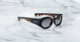 A pair of Jacques Marie Mage Gia black color, thick-framed sunglasses with grey lenses sits on a white marble surface with grey veins, with faint brand details visible on the inside of the left lens.