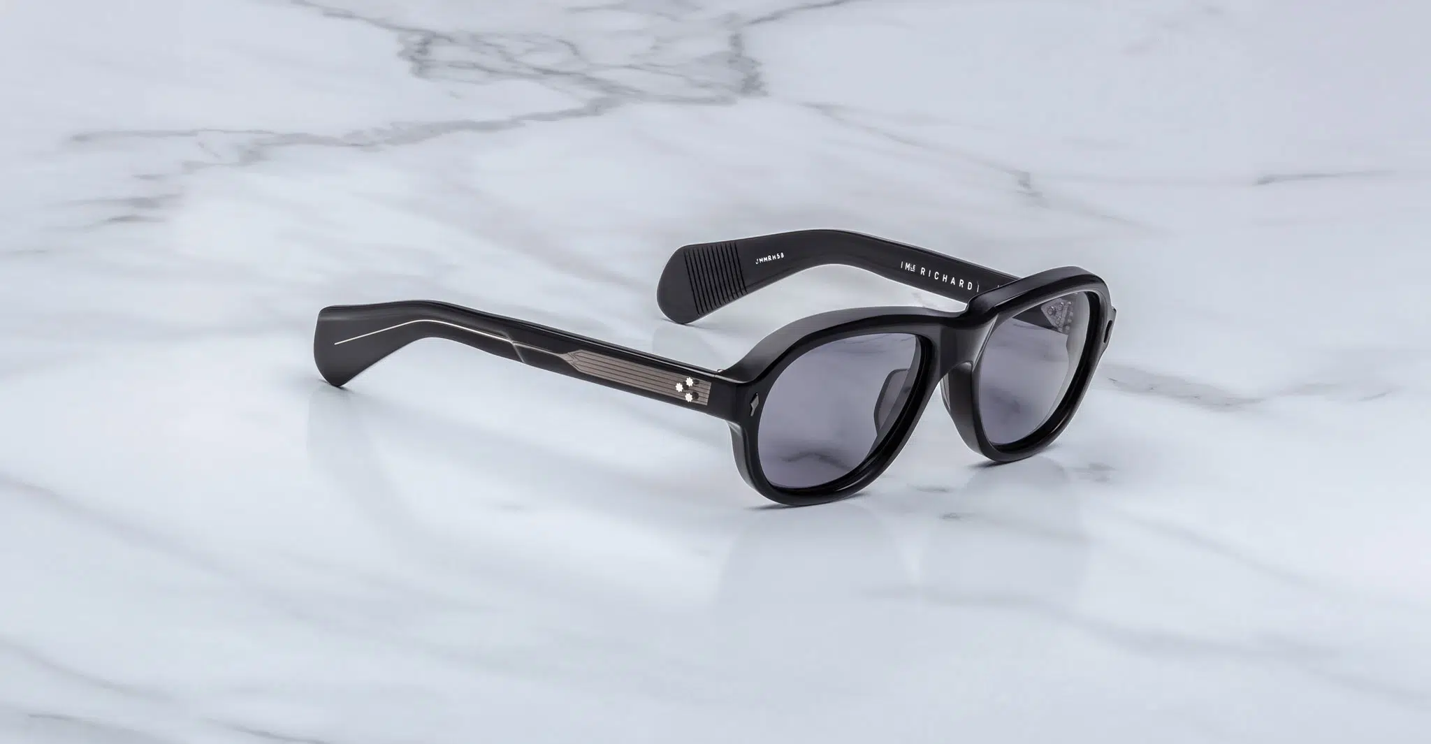 Jacques Marie Mage Richard black sunglasses with dark lenses and thick frames rest on a white marble surface, with the brand name visible on the inside of one temple arm.