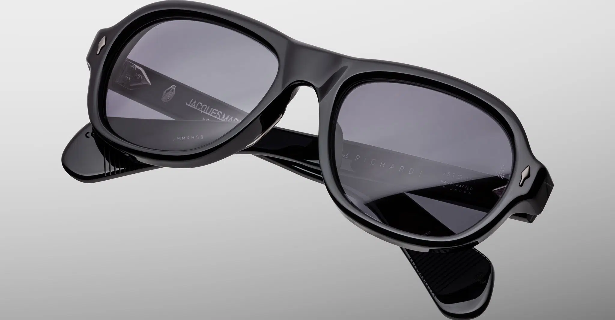 The Jacques Marie Mage Richard sunglasses feature thick, glossy black frames with dark lenses and visible brand markings on the arms, displayed against a light gray background.