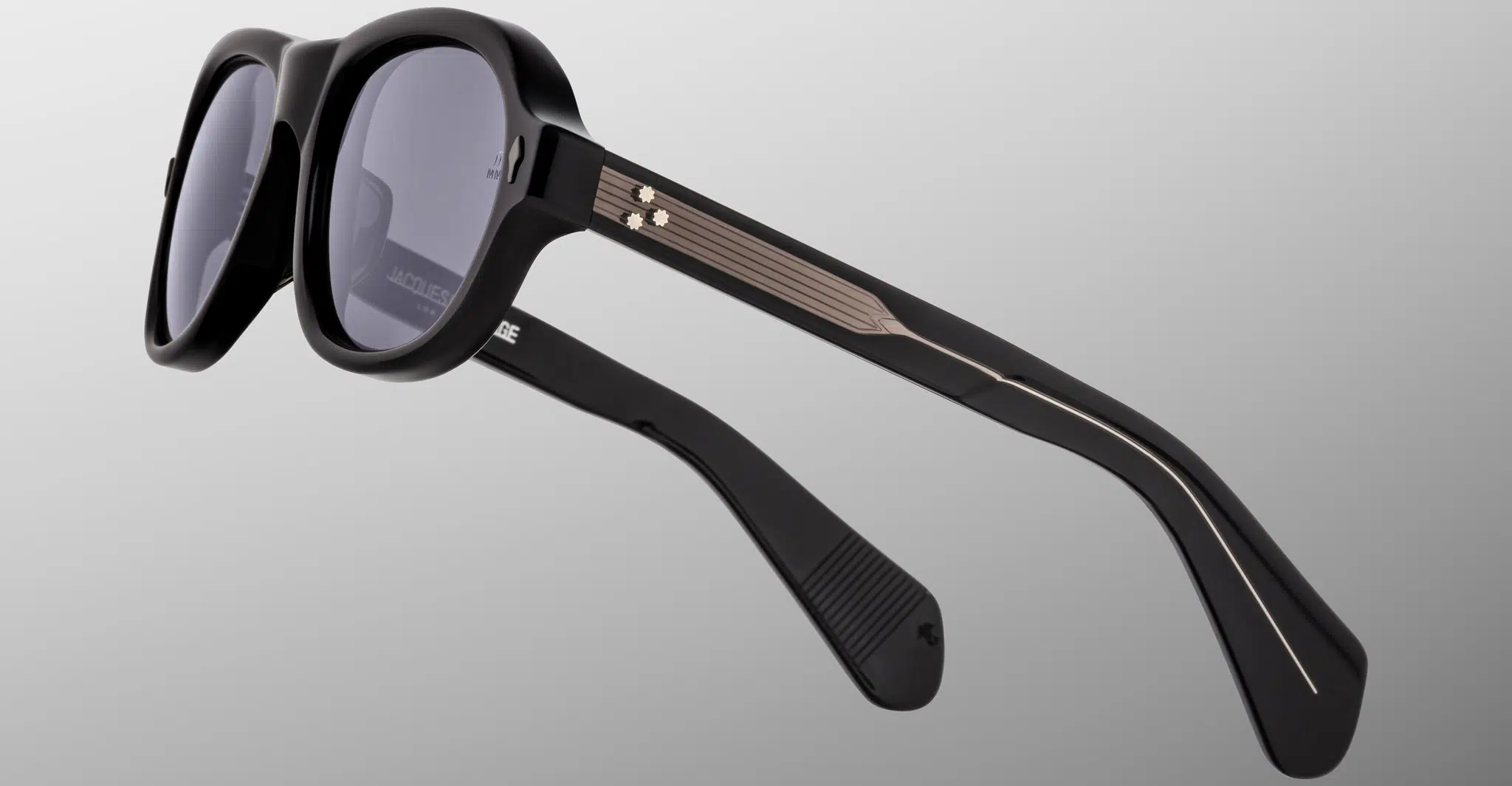 The Jacques Marie Mage Richard sunglasses feature thick black frames, dark lenses, and metallic gold accents with lined temples, displayed on a light gray background.