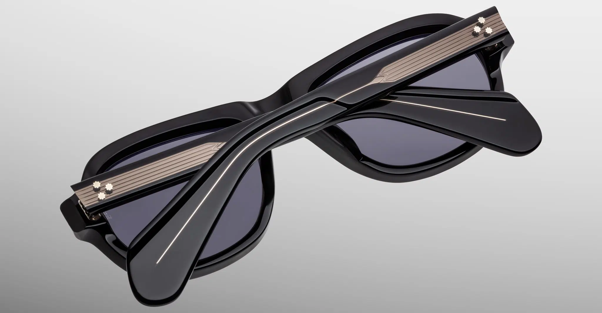 The Jacques Marie Mage Richard sunglasses are stylish black frames with crossed arms over dark lenses, metallic accents near the hinges, set against a light gradient background.