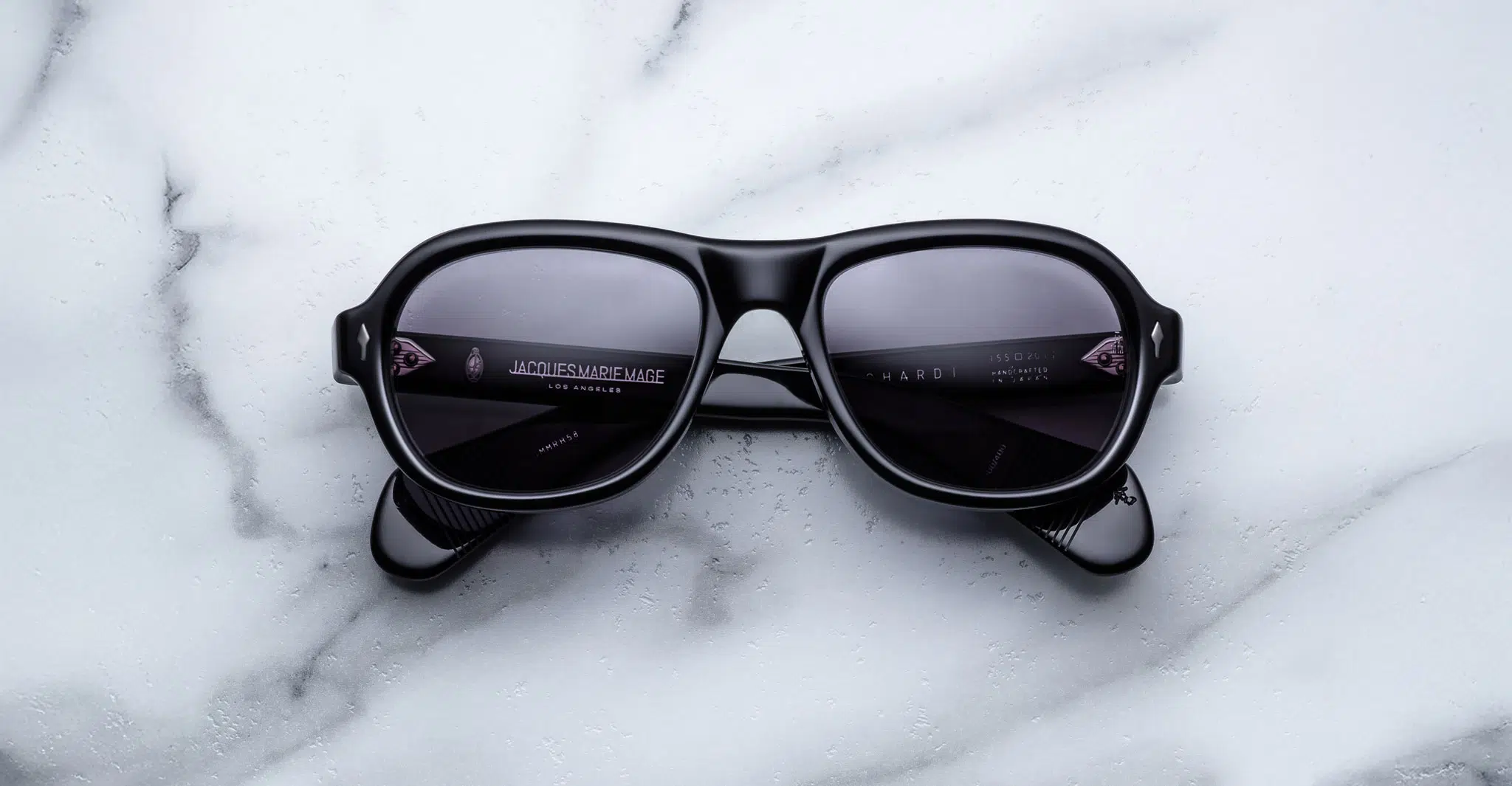 A pair of Jacques Marie Mage Richard black sunglasses with dark lenses sits centrally on a white marble surface, their arms neatly folded.