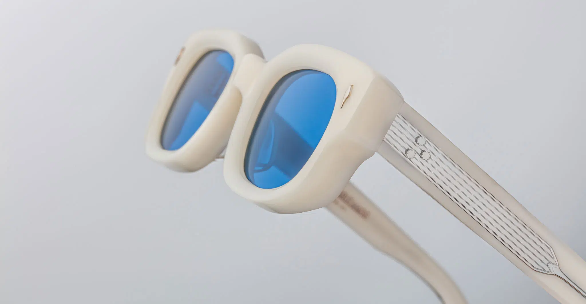 Close-up of Jacques Marie Mage Whiskeyclone sunglasses with thick cream frames, blue-tinted lenses, silver striped arms, and a small decorative detail, set against a light gray background.