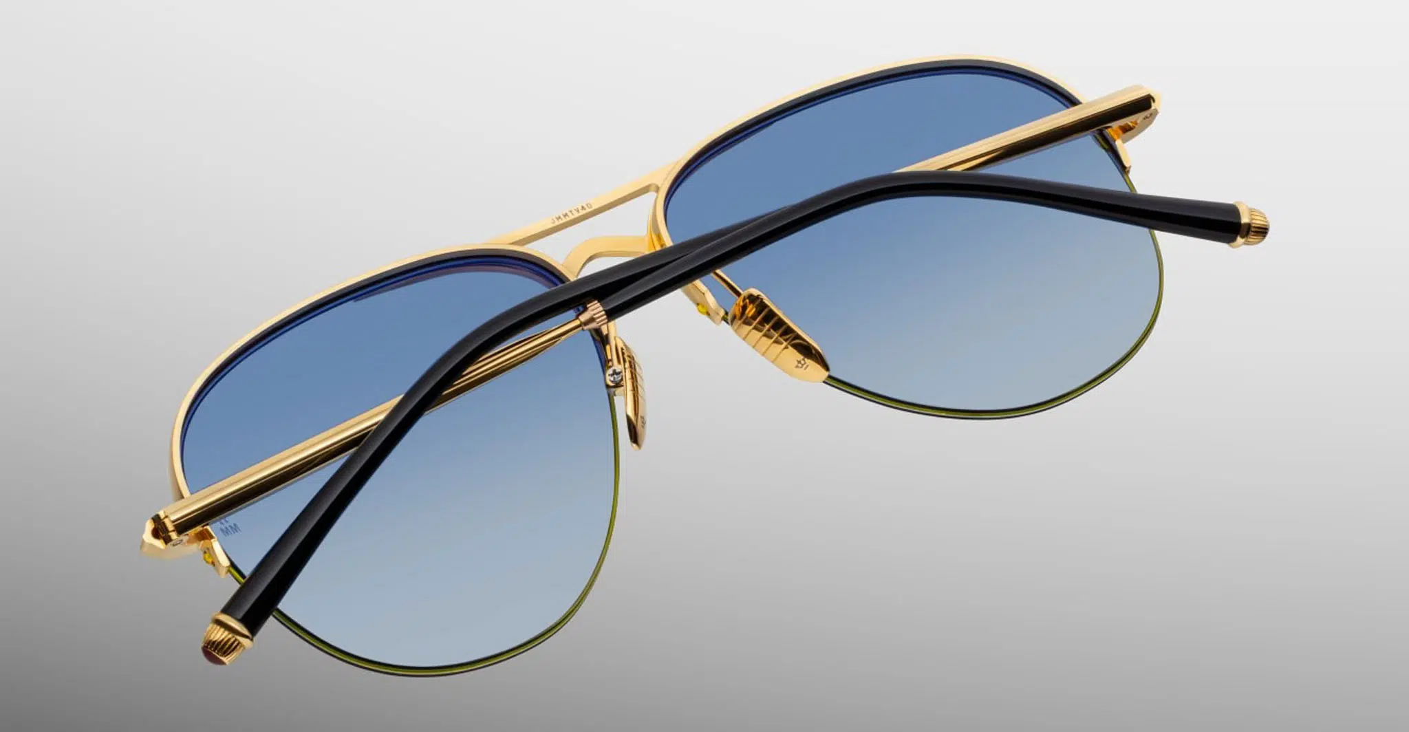 The Jacques Marie Mage Amelia sunglasses, featuring gold aviator frames and blue gradient lenses with black arms crossed over the lenses, are displayed on a light gray background.