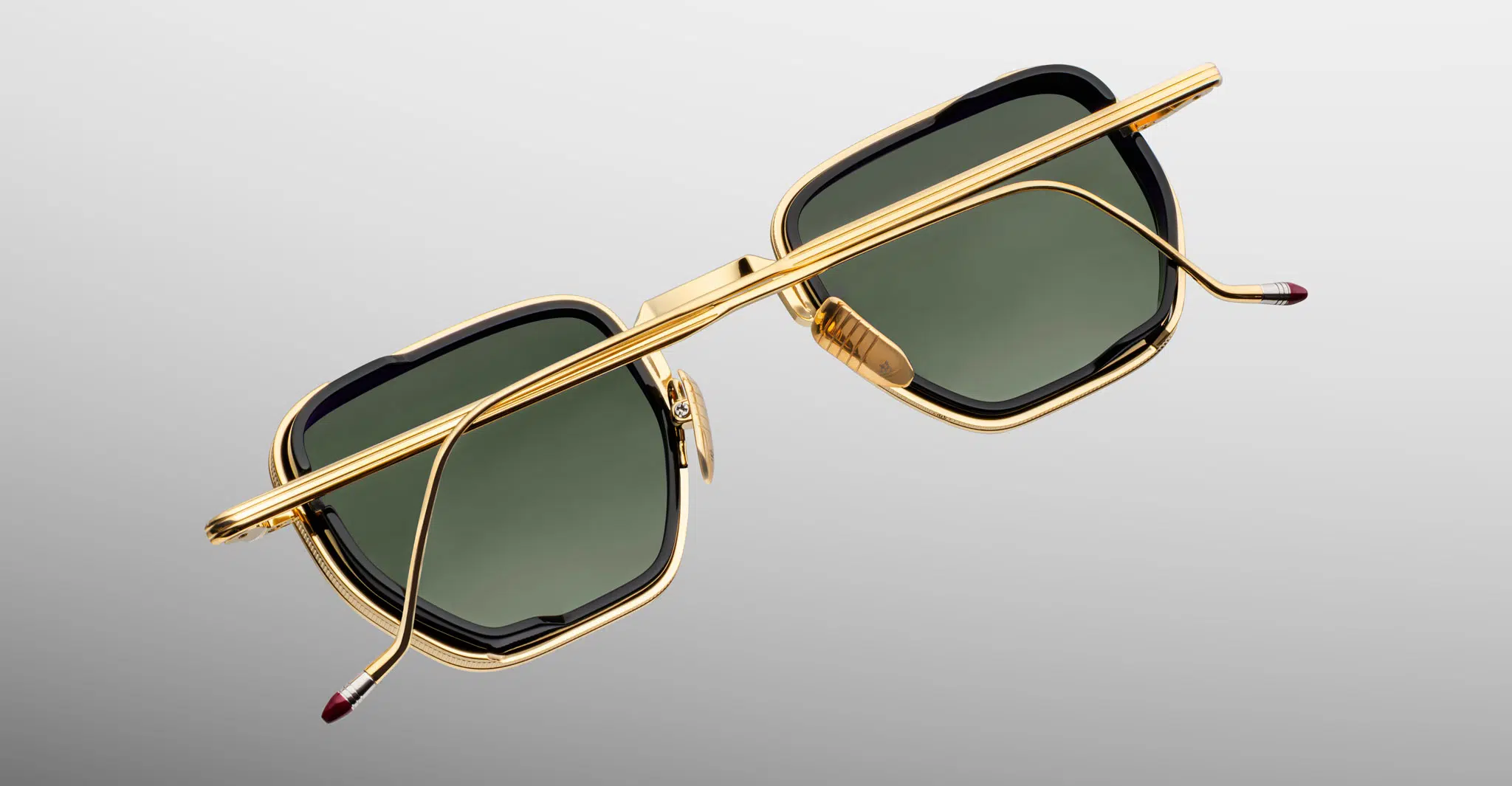 Jacques-Marie-Mage-Atkins-Musket-JMMTK-12M-Detail3-Dan-Deutsch The Jacques Marie Mage Atkins sunglasses, featuring gold rectangular frames and dark green lenses, are displayed from above on a light gray gradient, with their temples crossed over the lenses.