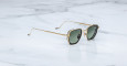 Jacques Marie Mage Atkins sunglasses with gold metal square frames and dark green lenses rest angled on a white marble surface, displaying both the side and front views.