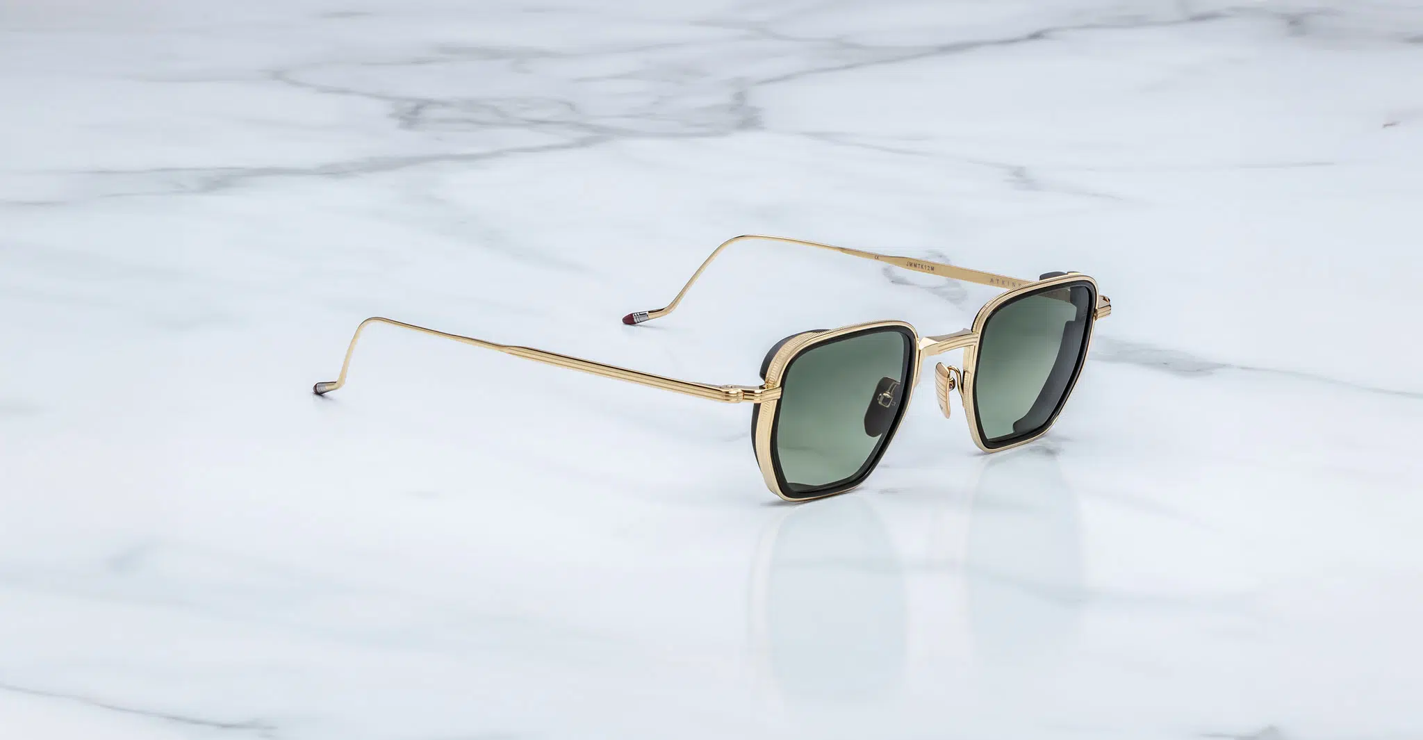 Jacques-Marie-Mage-Atkins-Musket-JMMTK-12M-Profile-Dan-Deutsch Jacques Marie Mage Atkins sunglasses with gold metal square frames and dark green lenses rest angled on a white marble surface, displaying both the side and front views.