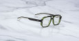 A pair of Jacques Marie Mage Evans eyeglasses with thick green and black rectangular frames and clear lenses rests on a white marble surface.