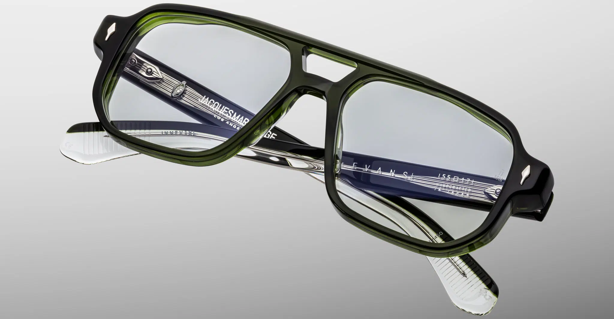 The Jacques Marie Mage Evans eyeglasses feature rectangular green transparent frames and clear lenses, with black and white patterned temples displaying branding. Shown against a light gray background.