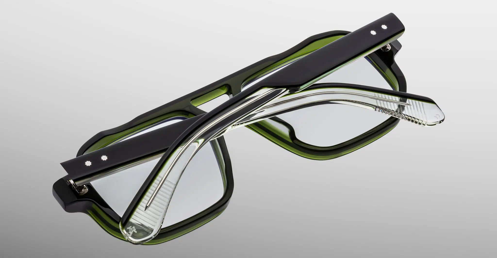 The Jacques Marie Mage Evans eyeglasses feature thick, rectangular green frames with black accents, displayed upside down with folded arms over the lenses against a light gray background.