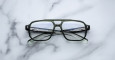 A pair of Jacques Marie Mage Evans rectangular eyeglasses with black and transparent frames lies on a white marble surface, arms crossed and text visible inside the left arm.