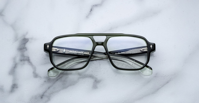 A pair of Jacques Marie Mage Evans rectangular eyeglasses with black and transparent frames lies on a white marble surface, arms crossed and text visible inside the left arm.
