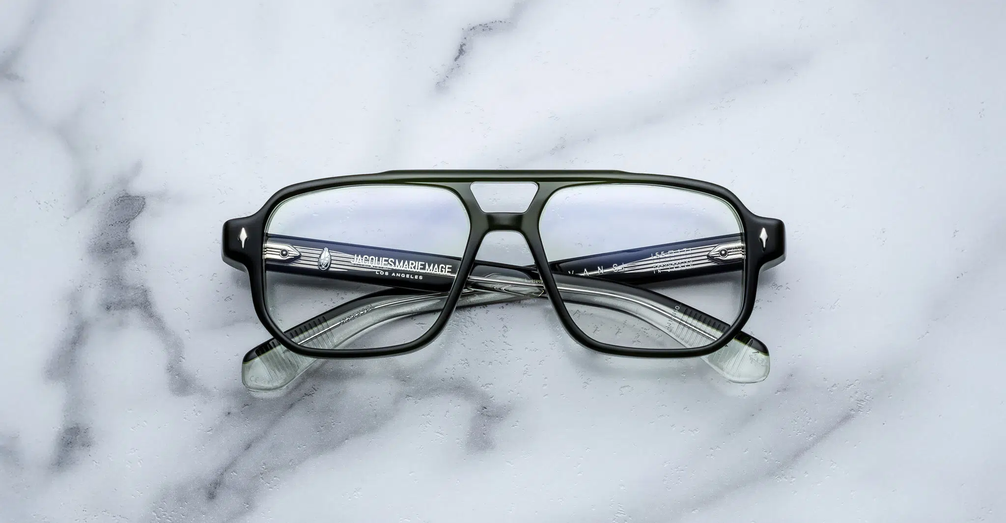 A pair of Jacques Marie Mage Evans rectangular eyeglasses with black and transparent frames lies on a white marble surface, arms crossed and text visible inside the left arm.