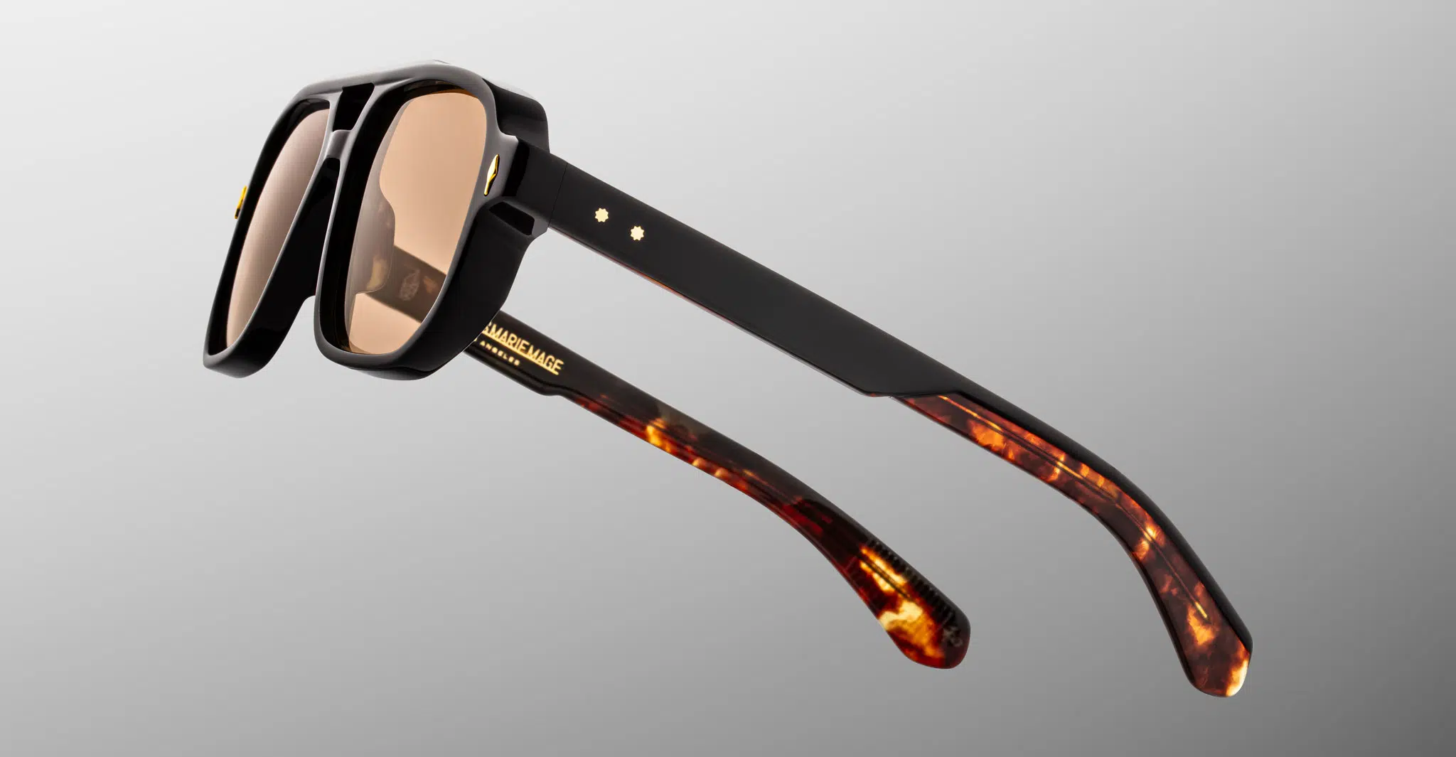 The Jacques Marie Mage Evans sunglasses feature thick black frames, brown tinted lenses, and glossy black arms that blend into a tortoiseshell pattern at the ends, set against a smooth grey gradient background.