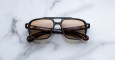 The Jacques Marie Mage Evans sunglasses feature brown-tinted, square lenses with a black and tortoiseshell frame, displayed on a white marble surface.