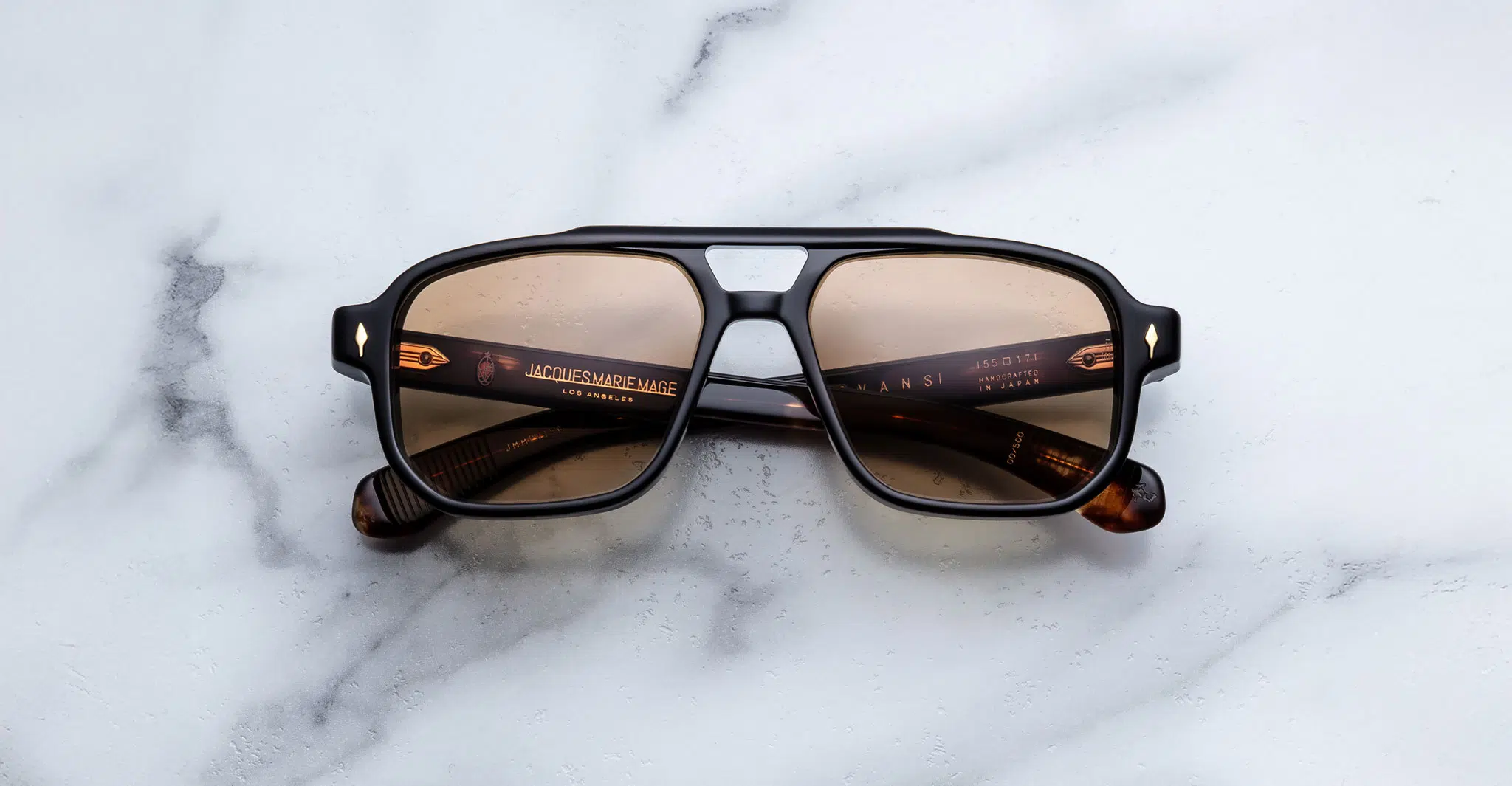 The Jacques Marie Mage Evans sunglasses feature brown-tinted, square lenses with a black and tortoiseshell frame, displayed on a white marble surface.