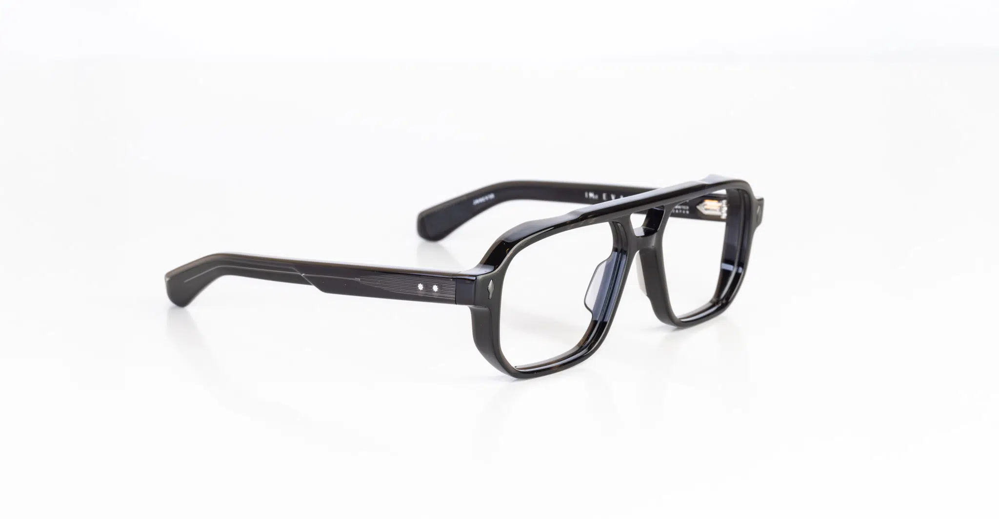 A pair of Jacques Marie Mage Evans black rectangular eyeglasses with thick frames and clear lenses rests on a white surface.