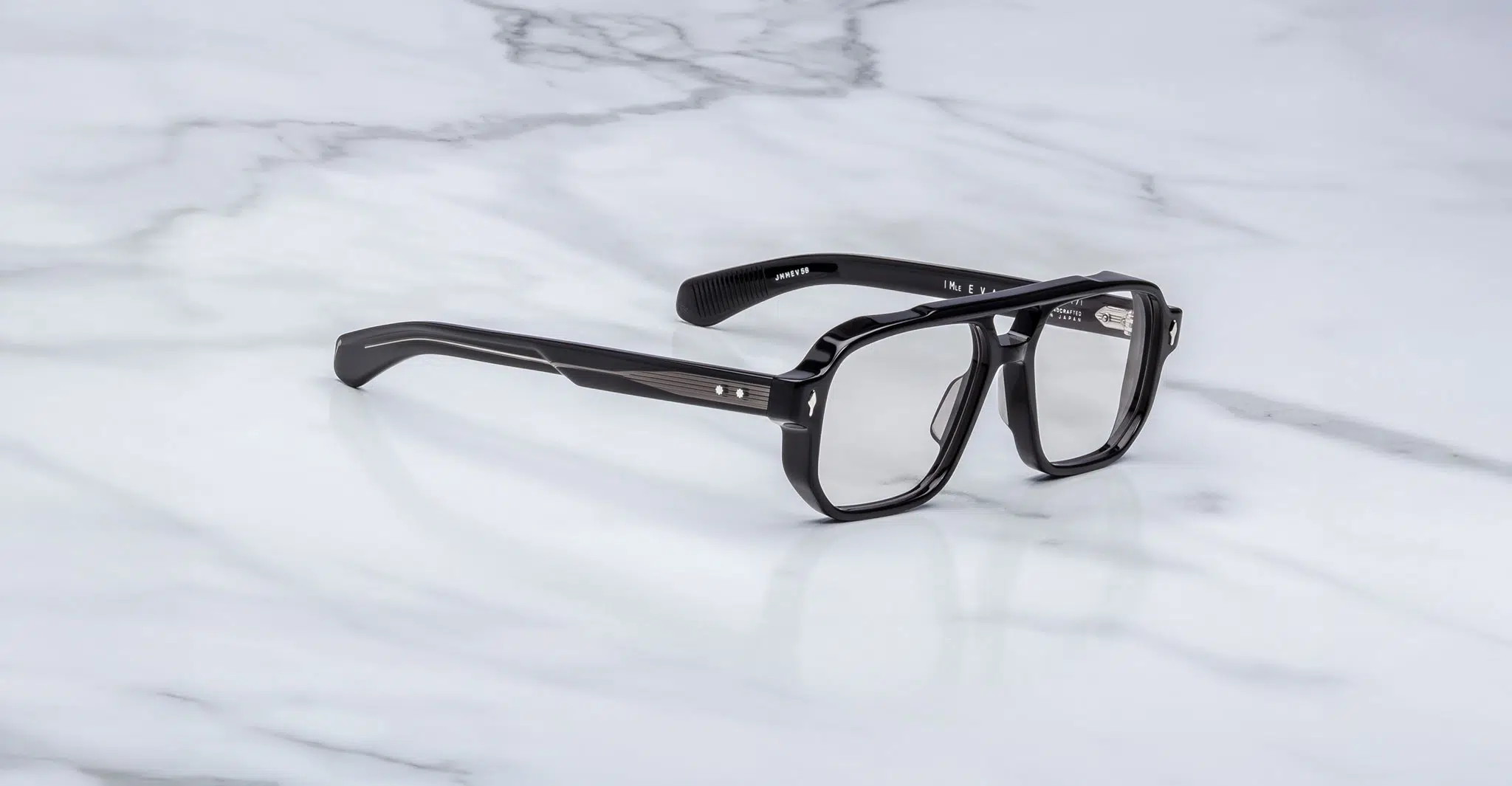 A pair of Jacques Marie Mage Evans black rectangular eyeglasses with clear lenses rests on a white marble surface.