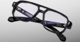 The Jacques Marie Mage Evans eyeglasses feature black, rectangular thick frames and clear lenses, shown on a gray background. Branding and model details in white text appear on the inner arm of the glasses.