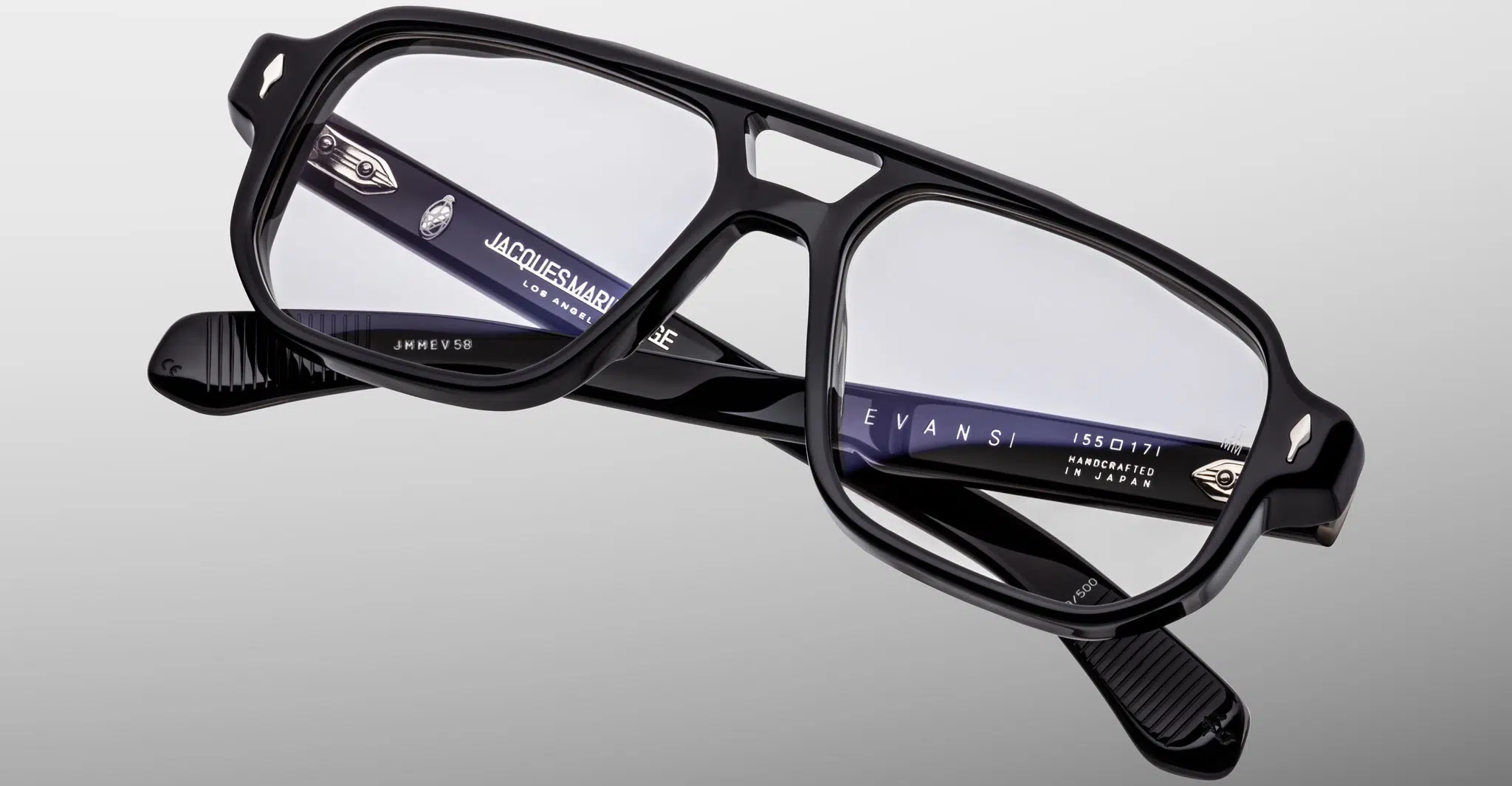 The Jacques Marie Mage Evans eyeglasses feature black, rectangular thick frames and clear lenses, shown on a gray background. Branding and model details in white text appear on the inner arm of the glasses.