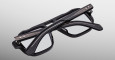 The Jacques Marie Mage Evans eyeglasses, black and rectangular with metallic accents, are shown folded with one temple arm crossed over the other on a plain light gray background.
