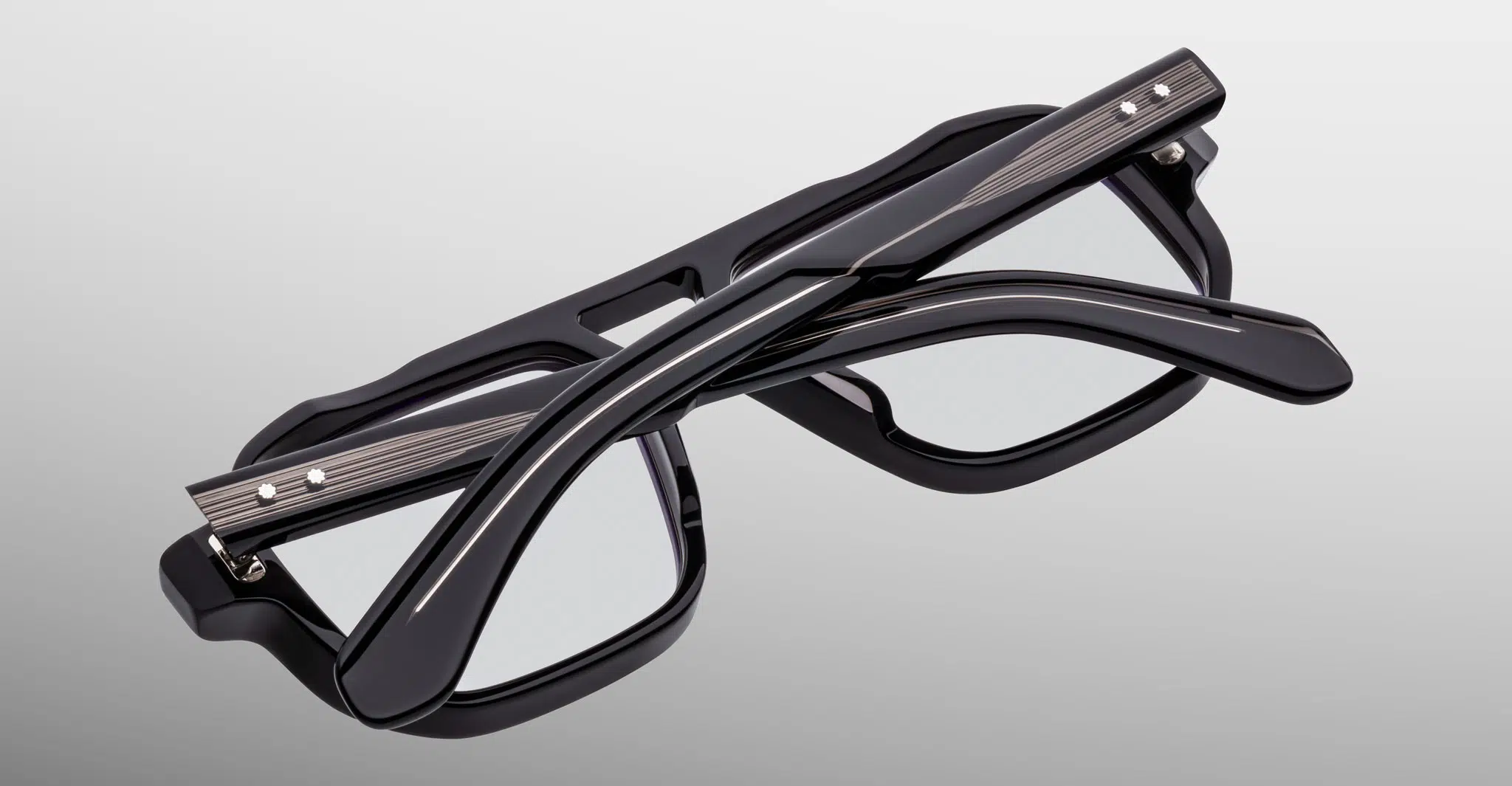 The Jacques Marie Mage Evans eyeglasses, black and rectangular with metallic accents, are shown folded with one temple arm crossed over the other on a plain light gray background.
