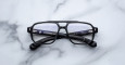 A pair of Jacques Marie Mage Evans black-rimmed eyeglasses with rectangular lenses rests on a white marble surface, showing brand and model details on the inner arms.