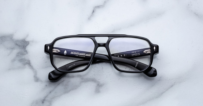 A pair of Jacques Marie Mage Evans black-rimmed eyeglasses with rectangular lenses rests on a white marble surface, showing brand and model details on the inner arms.