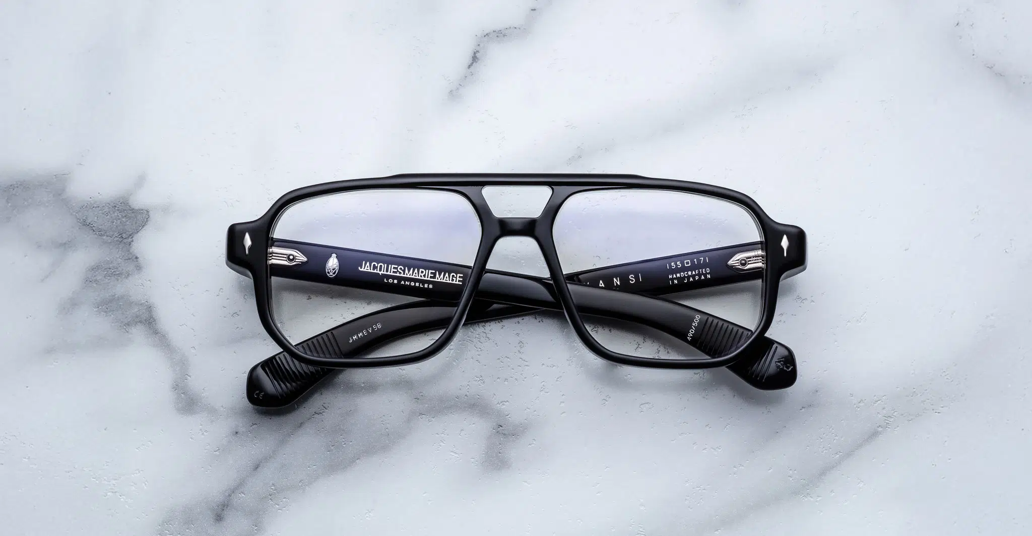 A pair of Jacques Marie Mage Evans black-rimmed eyeglasses with rectangular lenses rests on a white marble surface, showing brand and model details on the inner arms.