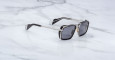 A pair of Jacques Marie Mage GT-R black square sunglasses with metal arms and dark lenses rests on a white marble surface.