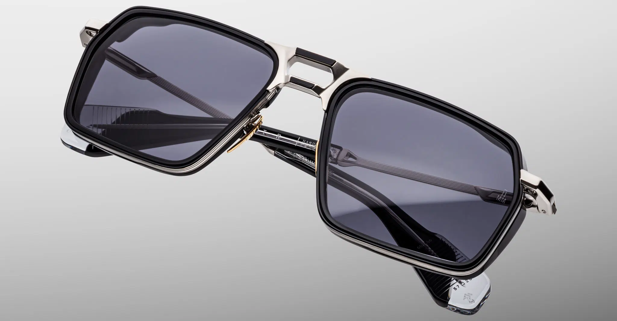 The Jacques Marie Mage GT-R features rectangular black frames, dark tinted lenses, and metallic accents on the bridge and temples, showcased against a neutral gradient background.
