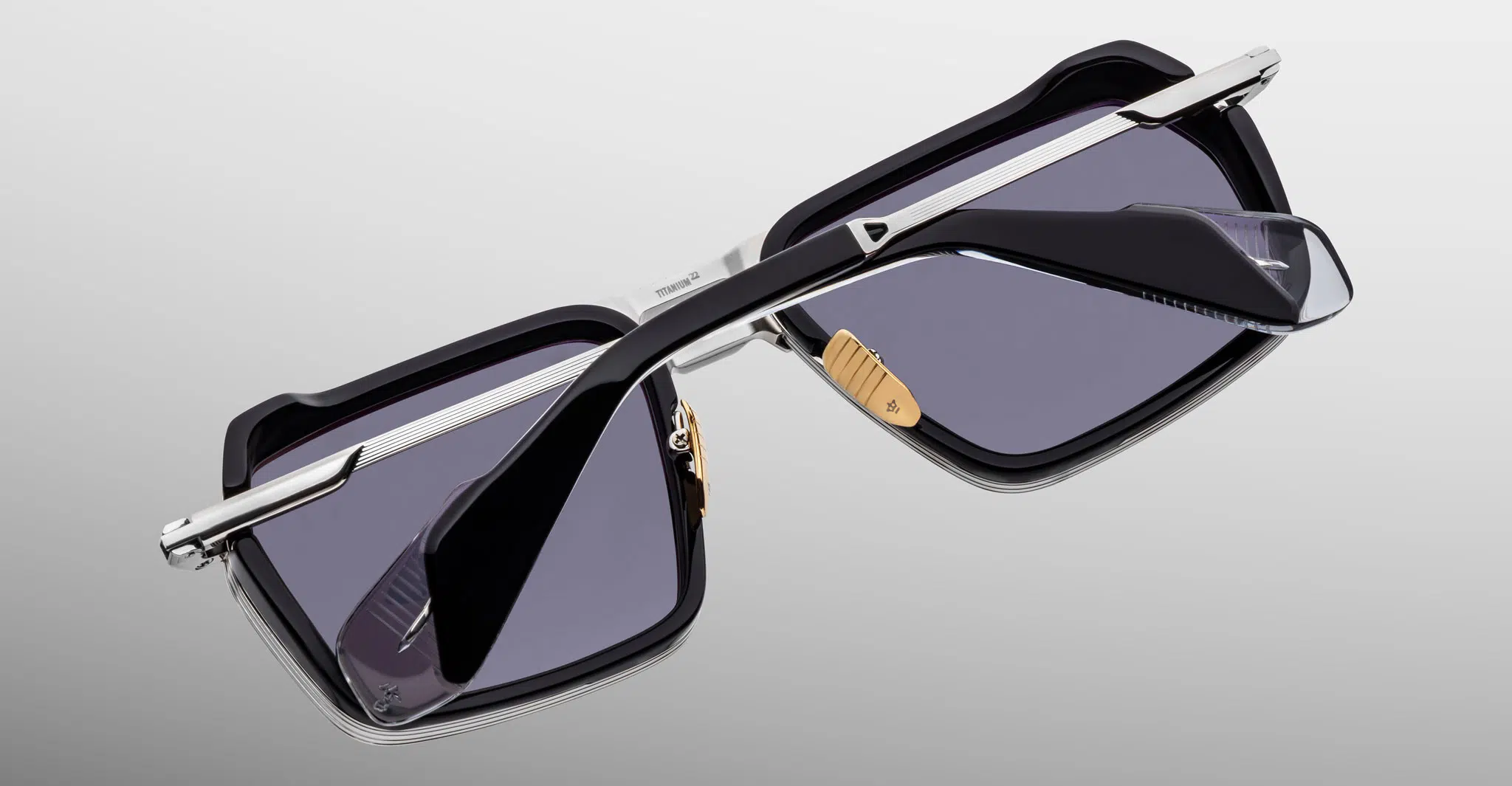 The Jacques Marie Mage GT-R sunglasses feature a black rectangular frame, silver metal arms, and dark tinted lenses, shown resting upside down on a neutral gradient background.