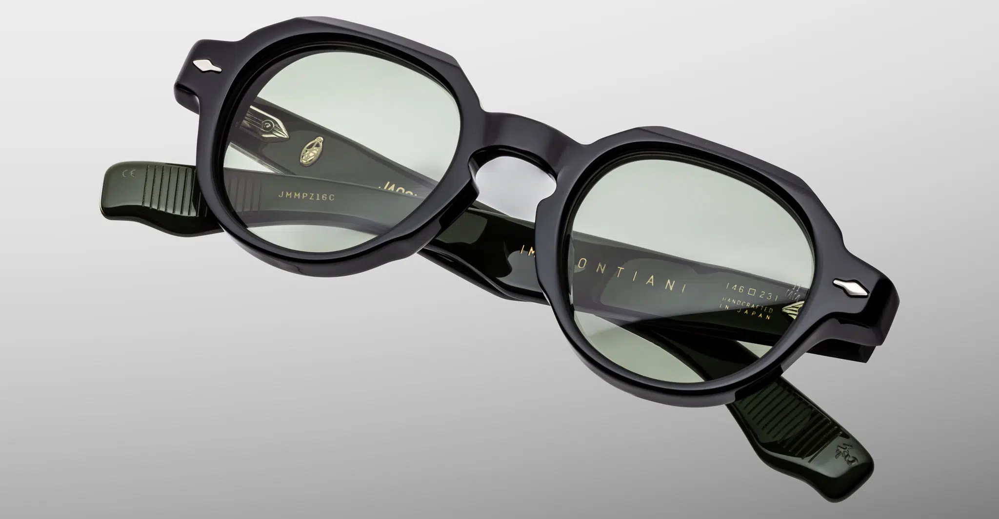 The Jacques Marie Mage Pontian sunglasses in black, featuring geometric frames and green-tinted lenses, are displayed on a plain light gray background. The brand name and details appear on the inside of the arms.