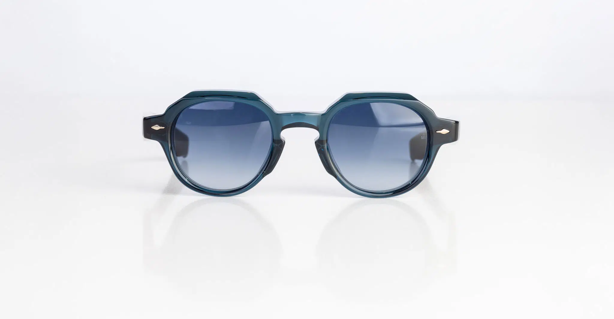 The Jacques Marie Mage Pontian sunglasses, featuring a round silhouette in dark teal with blue-tinted lenses, rest on a white surface against a plain white background.