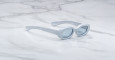 Jacques Marie Mage Besset sunglasses feature light blue, oval-shaped thick frames and lightly tinted lenses, elegantly displayed on a white marble surface.