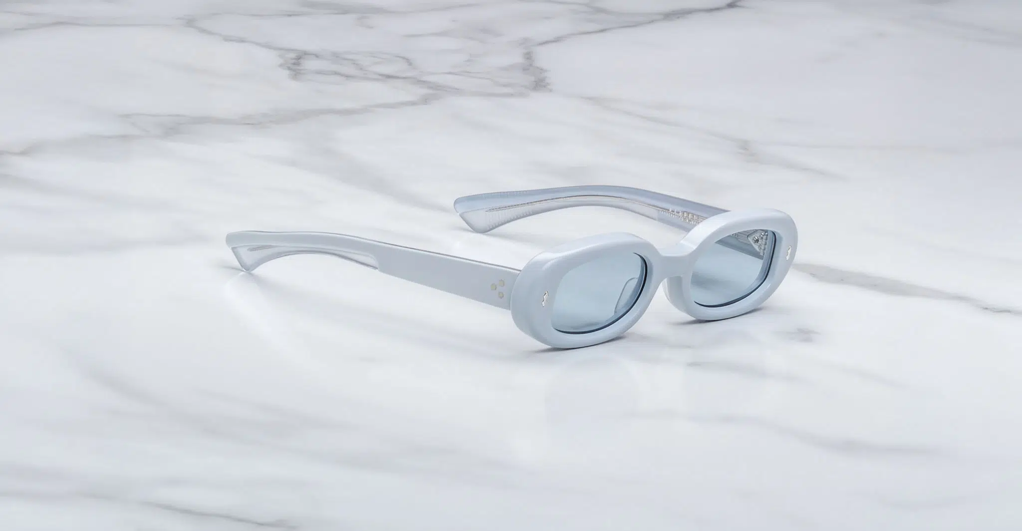 Jacques Marie Mage Besset sunglasses feature light blue, oval-shaped thick frames and lightly tinted lenses, elegantly displayed on a white marble surface.