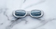 A pair of Jacques Marie Mage Besset white oval sunglasses with blue-tinted lenses rests on a white marble surface. The crossed temples and a metal railing reflected in the lenses add to their striking appearance.