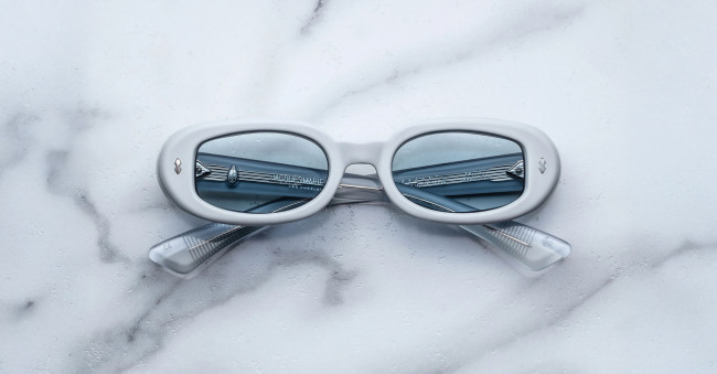 A pair of Jacques Marie Mage Besset white oval sunglasses with blue-tinted lenses rests on a white marble surface. The crossed temples and a metal railing reflected in the lenses add to their striking appearance.