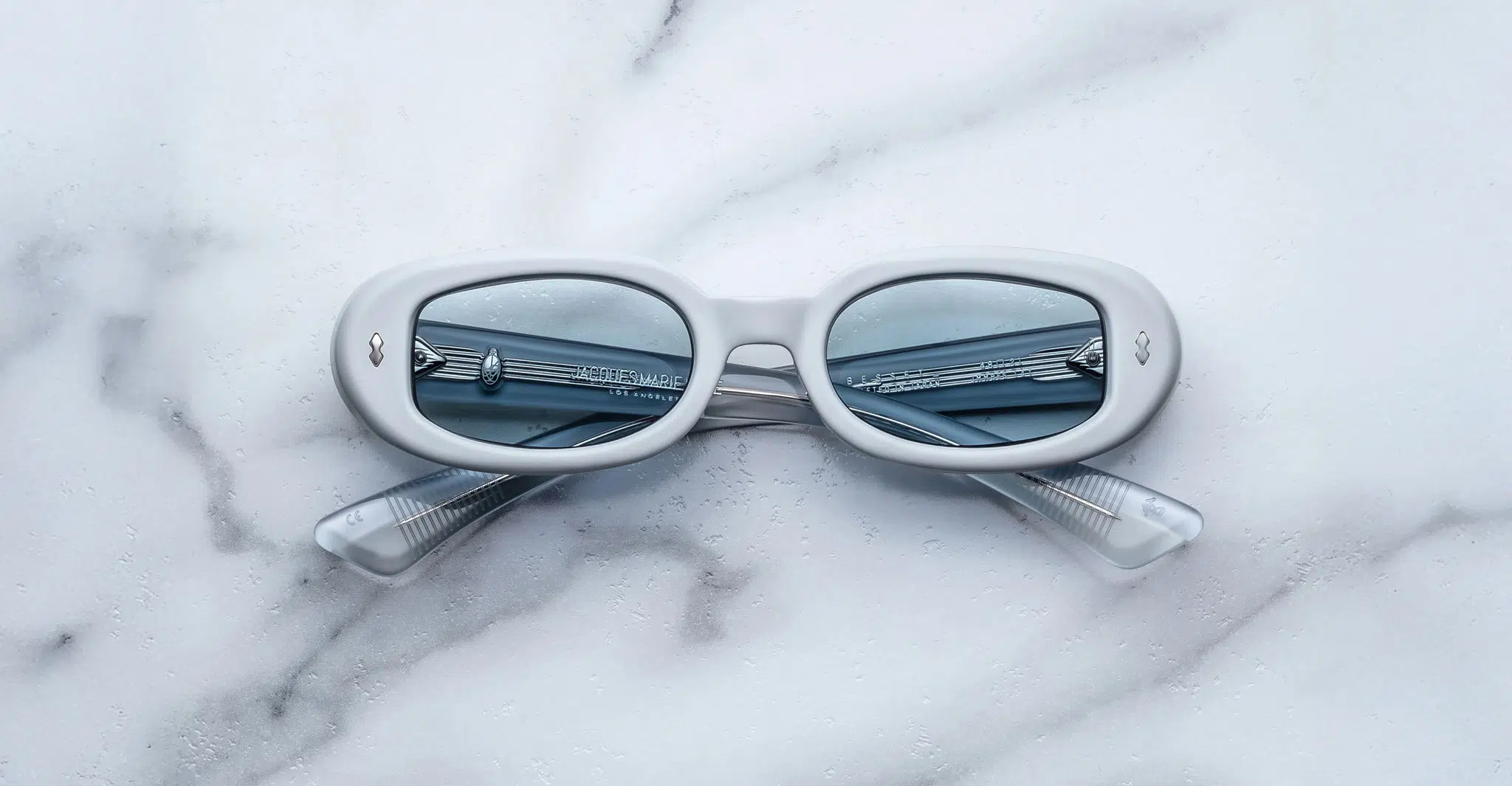 A pair of Jacques Marie Mage Besset white oval sunglasses with blue-tinted lenses rests on a white marble surface. The crossed temples and a metal railing reflected in the lenses add to their striking appearance.