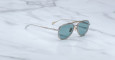 The Jacques Marie Mage Cougar sunglasses, featuring aviator-style thin metal frames and blue-green tinted lenses, rest on a white marble surface with gray veining.