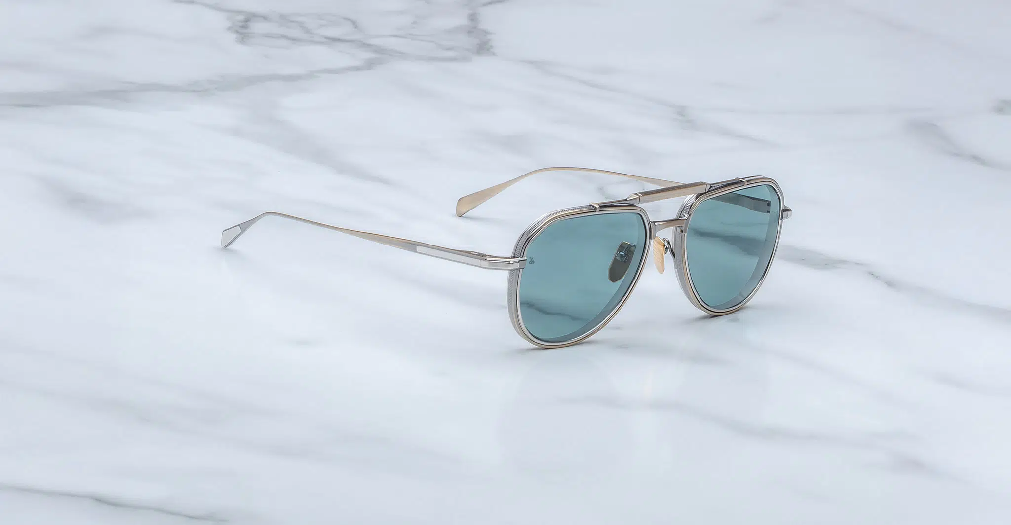 The Jacques Marie Mage Cougar sunglasses, featuring aviator-style thin metal frames and blue-green tinted lenses, rest on a white marble surface with gray veining.