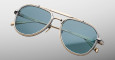 The Jacques Marie Mage Cougar sunglasses feature a gold metal aviator frame and blue-tinted lenses, displayed against a plain gray background.