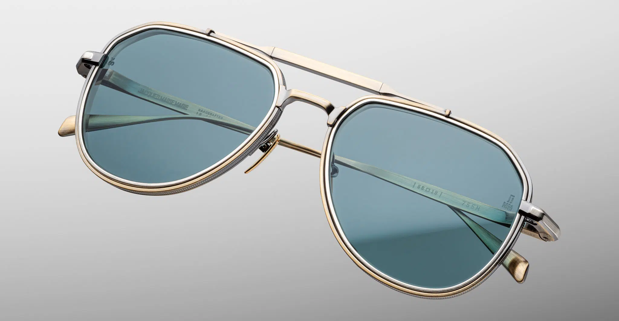 The Jacques Marie Mage Cougar sunglasses feature a gold metal aviator frame and blue-tinted lenses, displayed against a plain gray background.