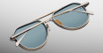 A close-up of folded Jacques Marie Mage Cougar sunglasses featuring blue-tinted lenses, metallic frames, and gold accents, displayed on a plain grey background.
