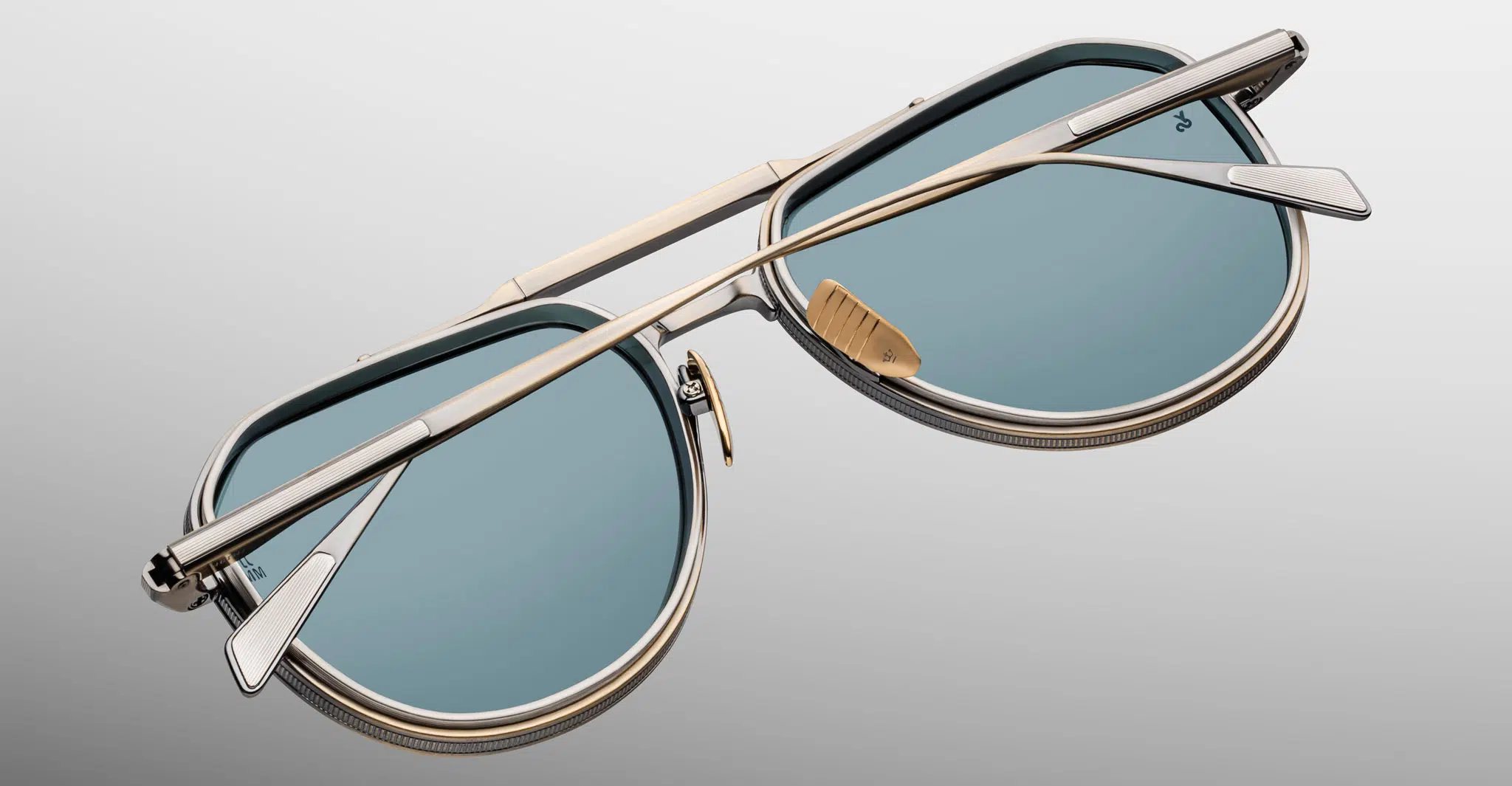 A close-up of folded Jacques Marie Mage Cougar sunglasses featuring blue-tinted lenses, metallic frames, and gold accents, displayed on a plain grey background.
